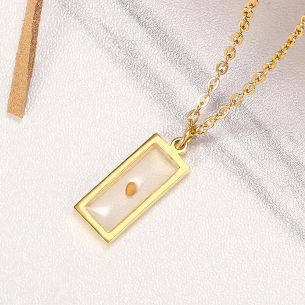 A gold rectangular pendant necklace with a clear front and a small amber bead suspended inside, displayed on a textured white surface.