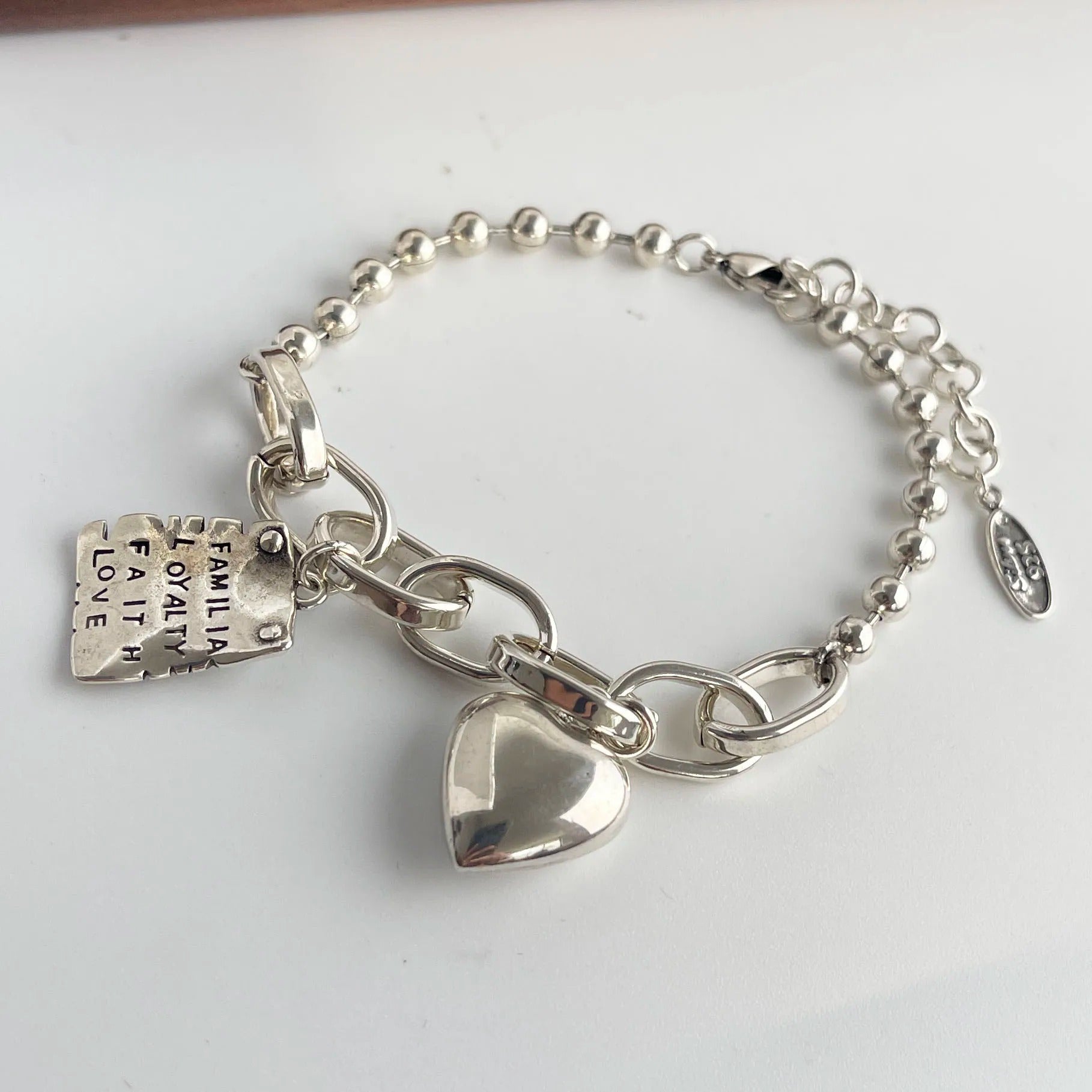 Silver bracelet with heart charm and engraved tag on a white background