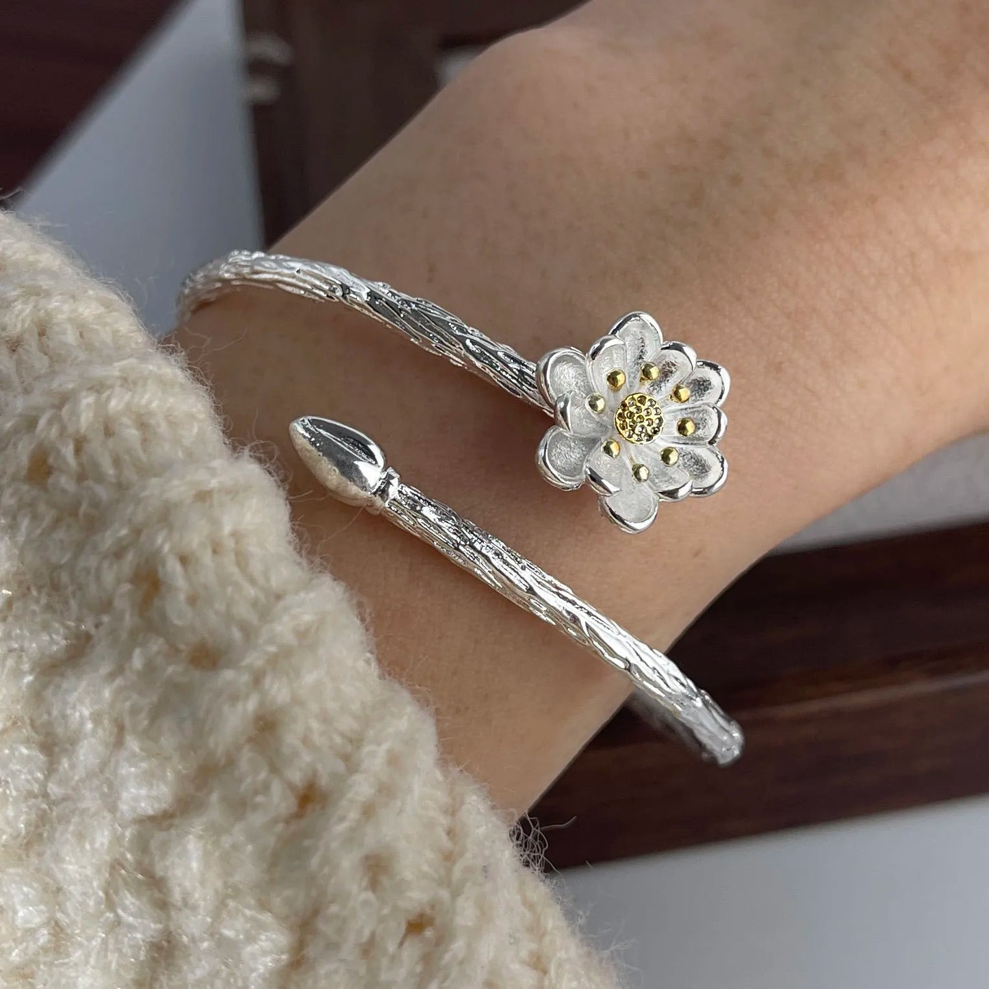 Silver bracelet with a flower charm on a wrist, wearing a beige sweater.