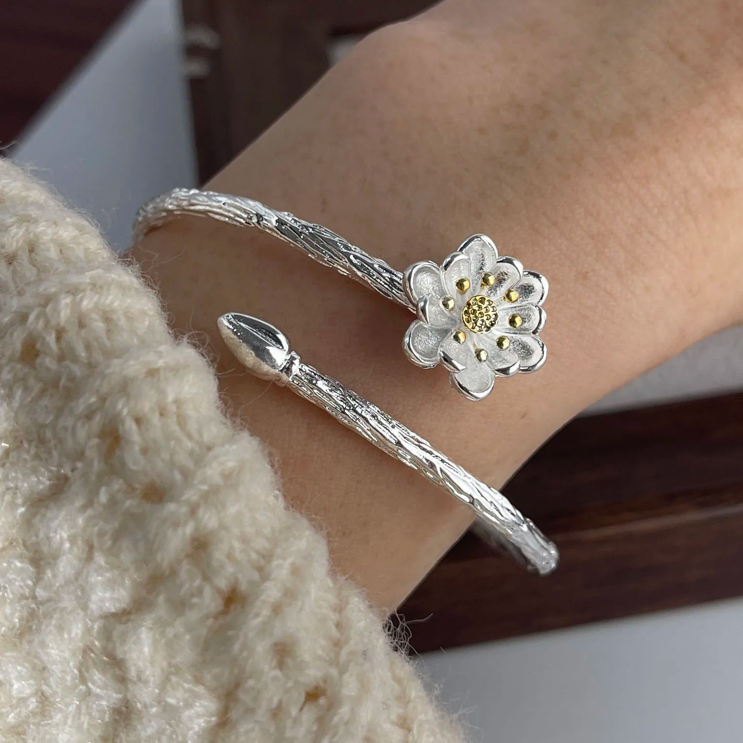 Silver bracelet with a flower charm on a wrist, wearing a beige sweater.