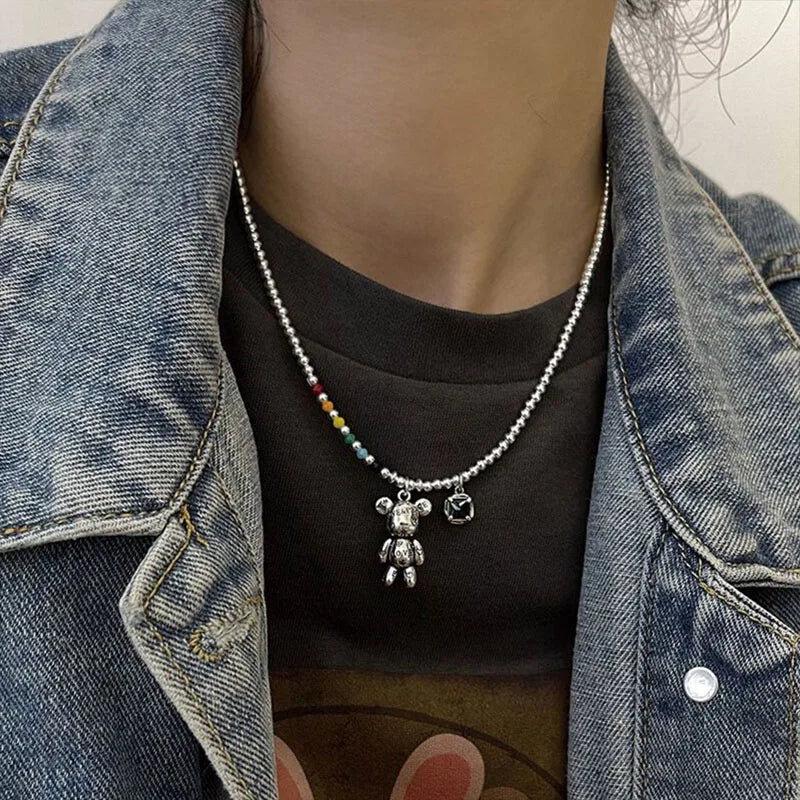 A close-up of a person wearing a silver beaded necklace with a teddy bear charm and a small round charm, styled over a black t-shirt and a blue denim jacket.