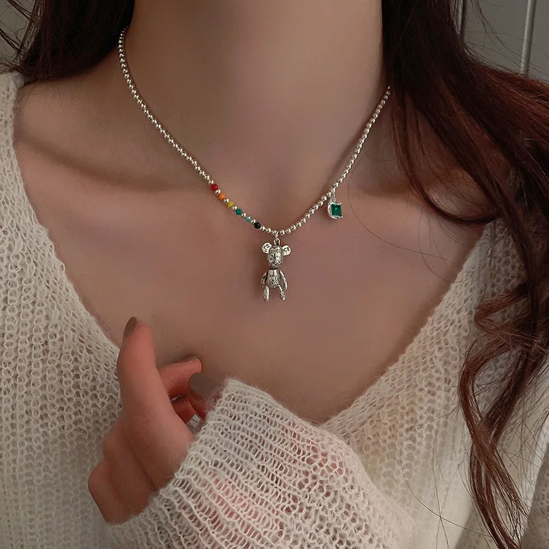 A close-up of a person wearing a delicate silver beaded necklace with a small teddy bear pendant, colorful accent beads, and a green square charm, paired with a cream knit V-neck sweater.