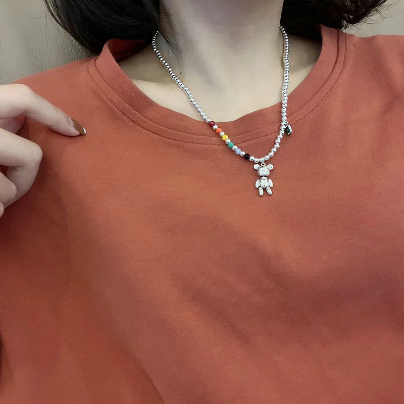 A close-up of a person wearing a silver beaded necklace with a small teddy bear pendant and a short rainbow bead section, paired with a rust-orange crewneck t-shirt.