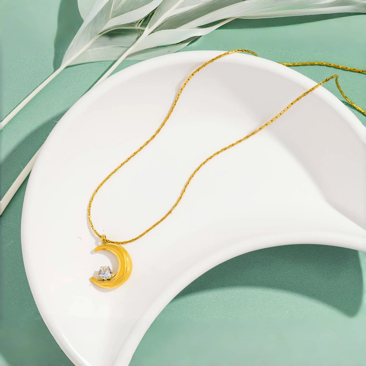 Gold necklace with a crescent moon pendant on a white dish with green leaves in the background