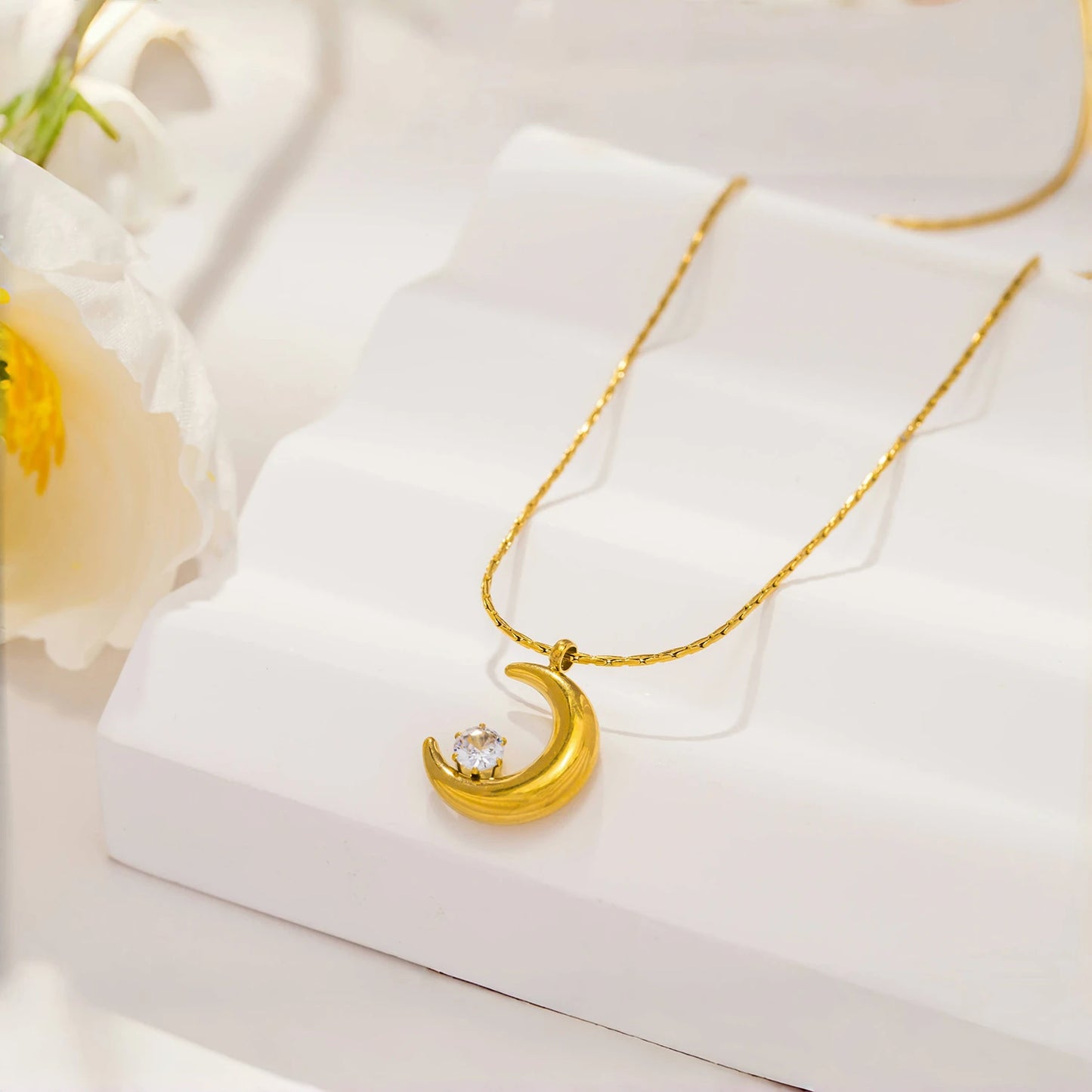 Gold necklace with a crescent moon pendant on a white surface with flowers.
