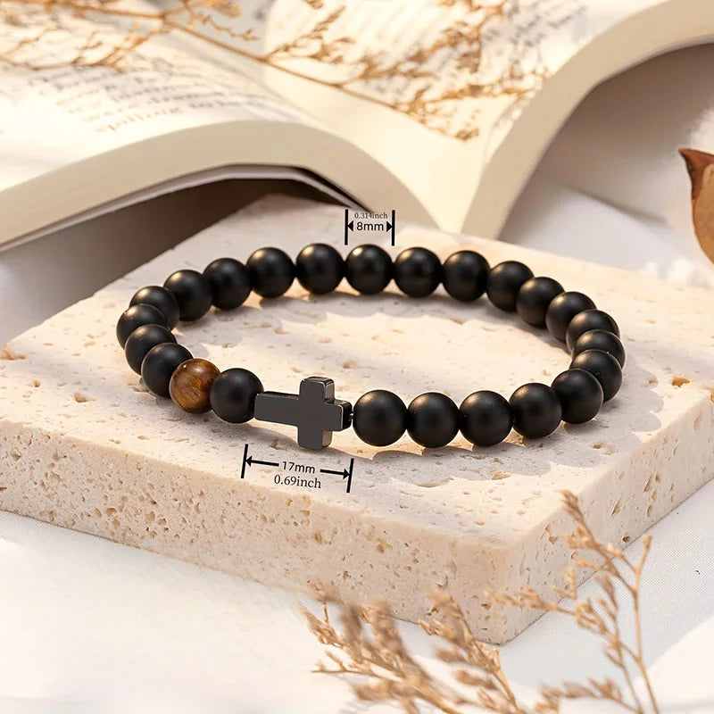 Black beaded bracelet with a cross charm on a stone surface with dried leaves.