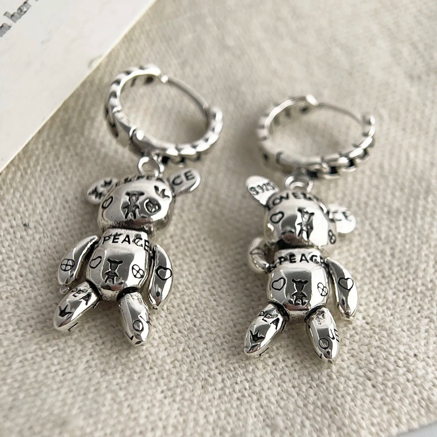 Pair of silver teddy bear keychains with 'Peace' text on a textured surface.