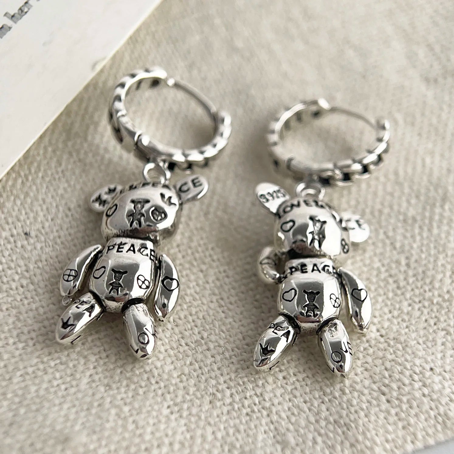 Pair of silver teddy bear keychains with 'Peace' text on a textured surface.