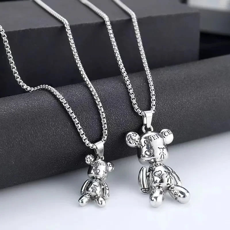 Two silver teddy bear pendant necklaces on matching chain, displayed on a dark gray cylindrical stand against a light gray background.