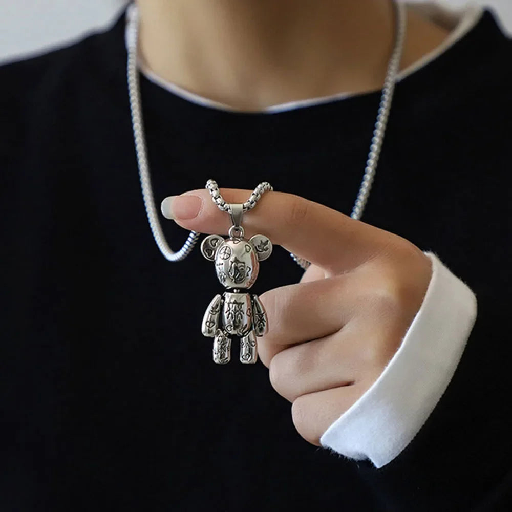 A person wearing a black top with a white collar and cuff, holding a silver bear pendant necklace in front of their chest. The necklace has a silver ball chain and a detailed teddy bear pendant with engraved patterns.
