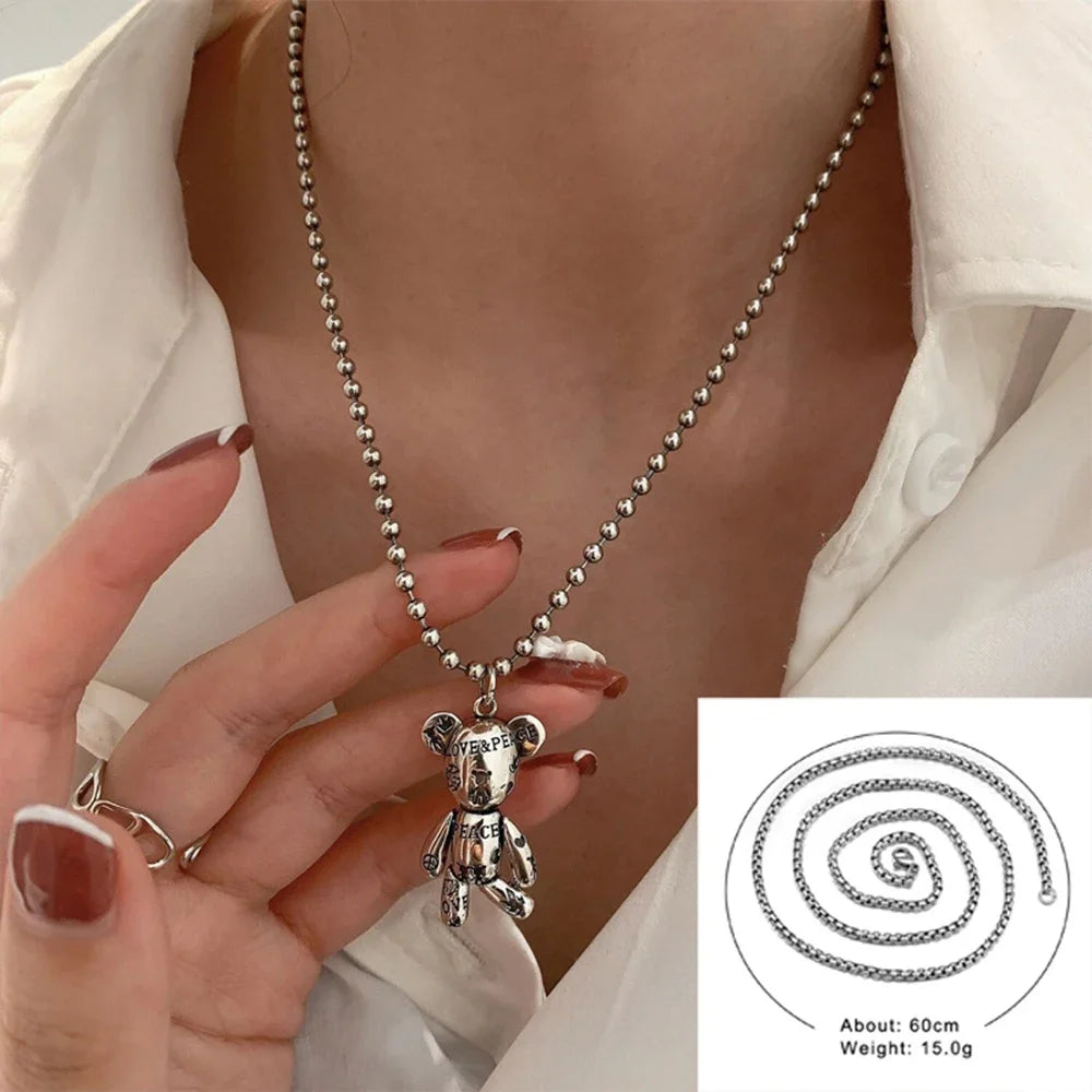A silver-toned beaded necklace with a teddy bear pendant worn by a person, shown with a hand holding part of the chain. A small inset in the bottom right shows the necklace coiled on a white background with text noting about 60 cm length and 15.