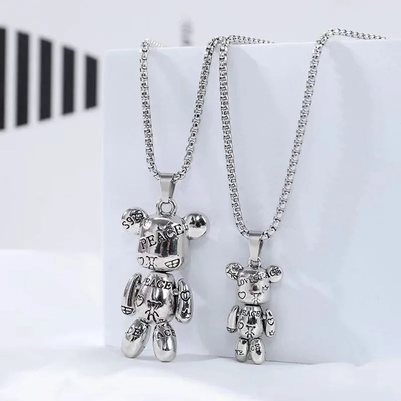 Two silver bear-shaped pendant necklaces on matching chains, displayed against a white block background. One pendant has 'PEACE' engraved on its face and arms, and the other has heart-shaped engravings.