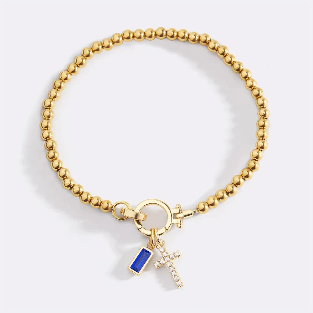 Gold bracelet with a blue charm and cross on a light gray background