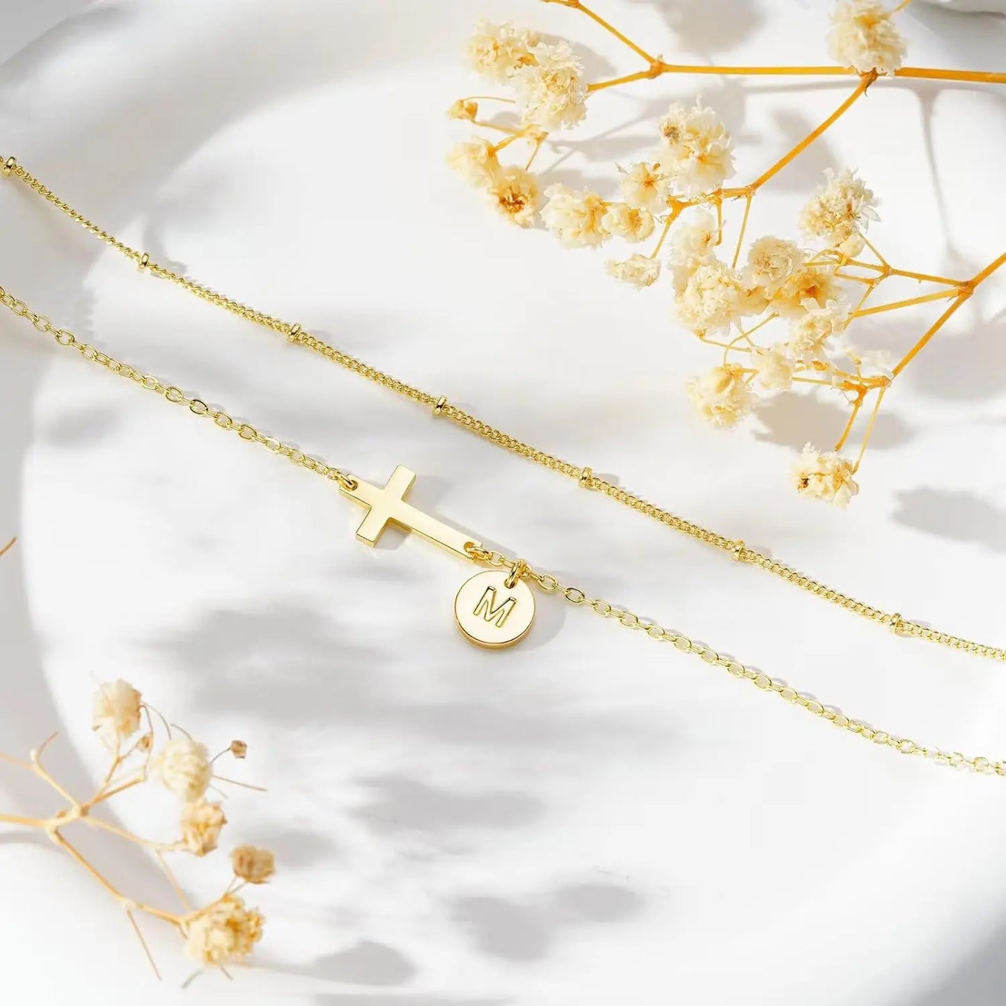 Gold necklaces with a cross and letter 'M' pendant on a white background with dried flowers.