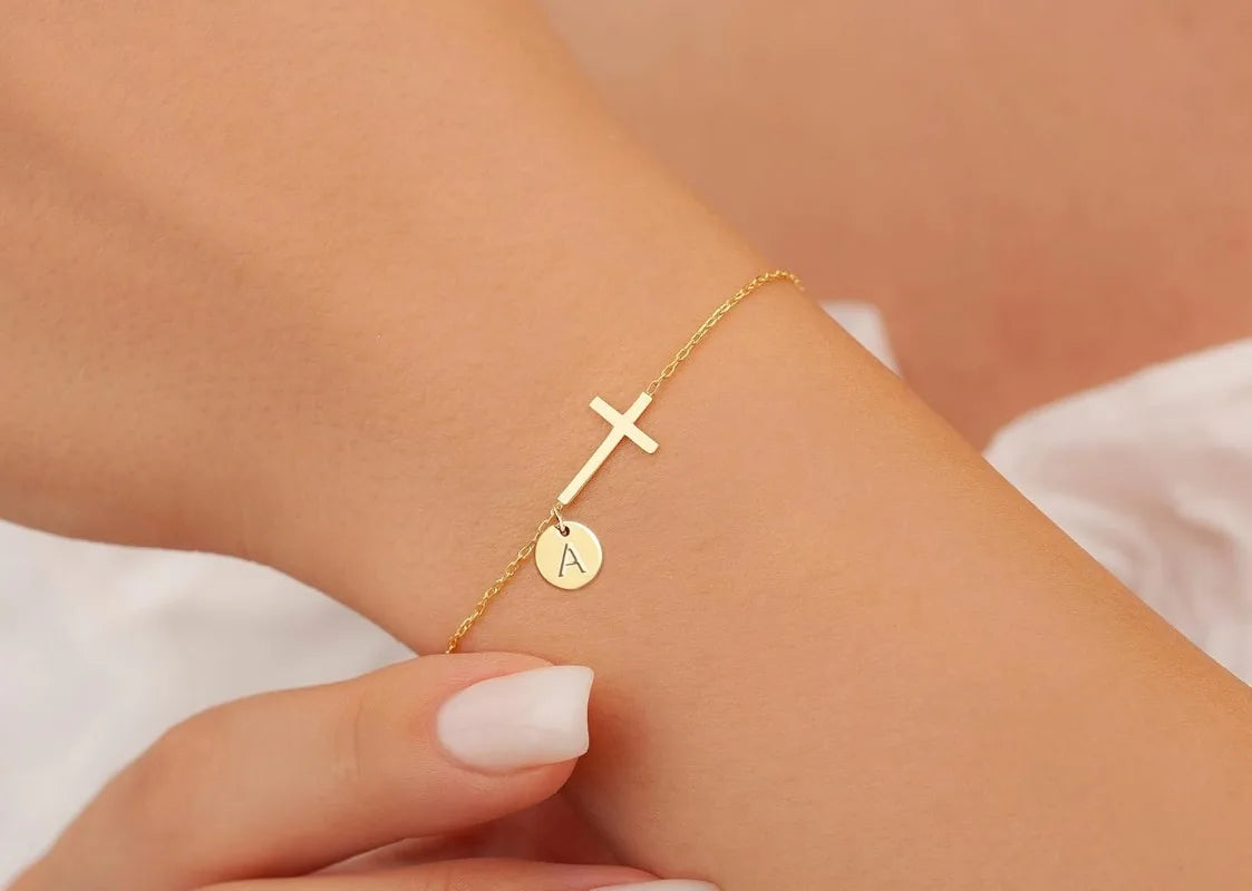 Gold bracelet with a cross and circular charm on a wrist against a neutral background