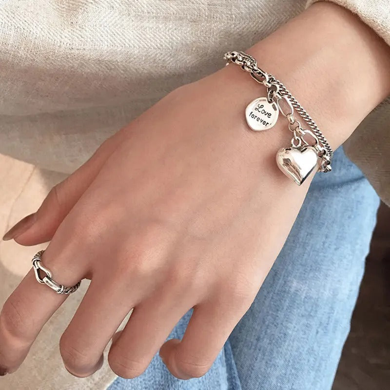 Hand wearing a silver bracelet with heart charm and ring on a neutral background