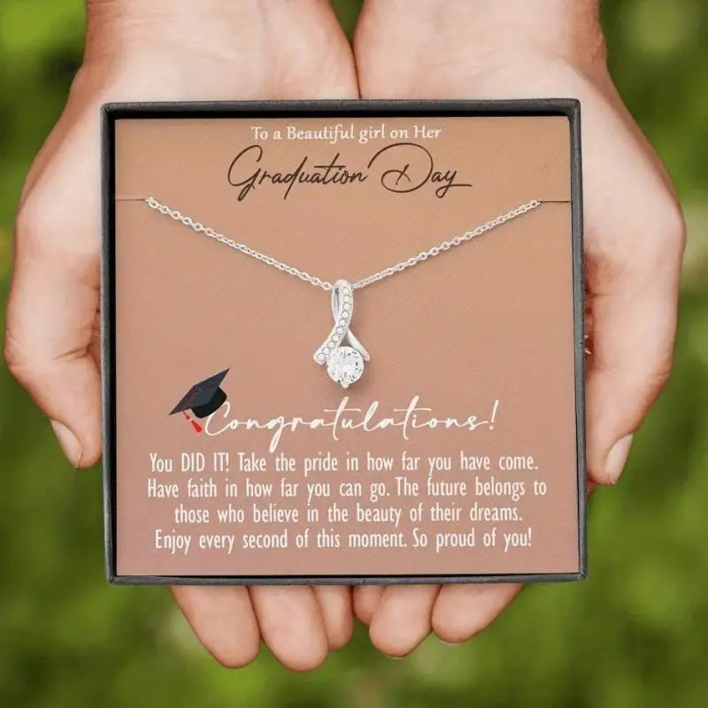 Graduation-themed necklace in a gift box held by hands with a green blurred background