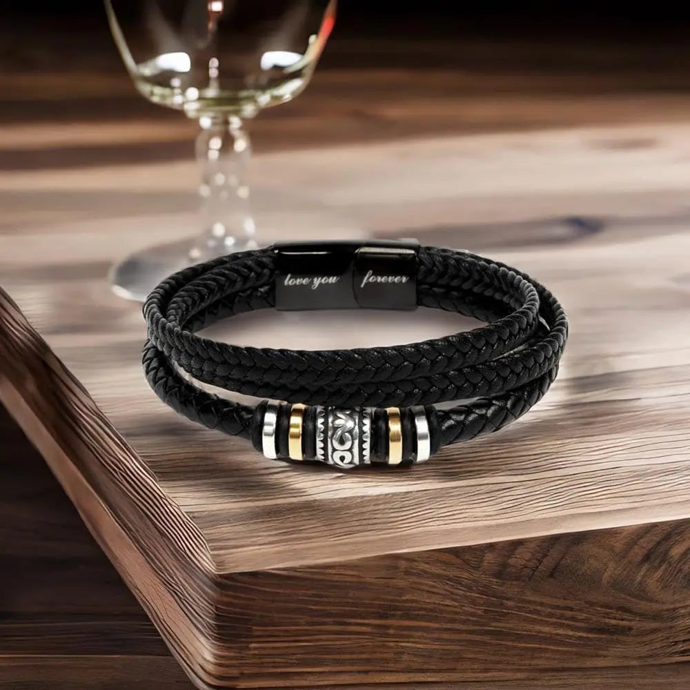 Black braided bracelet with metal accents on a wooden surface next to a glass of red wine.