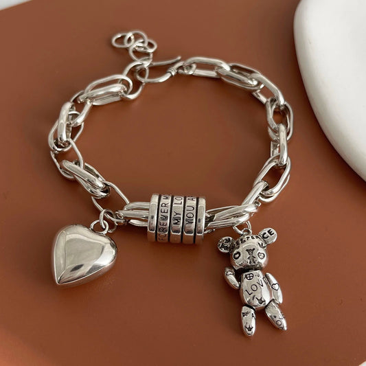 Silver chain bracelet with heart and teddy bear charms on a brown surface