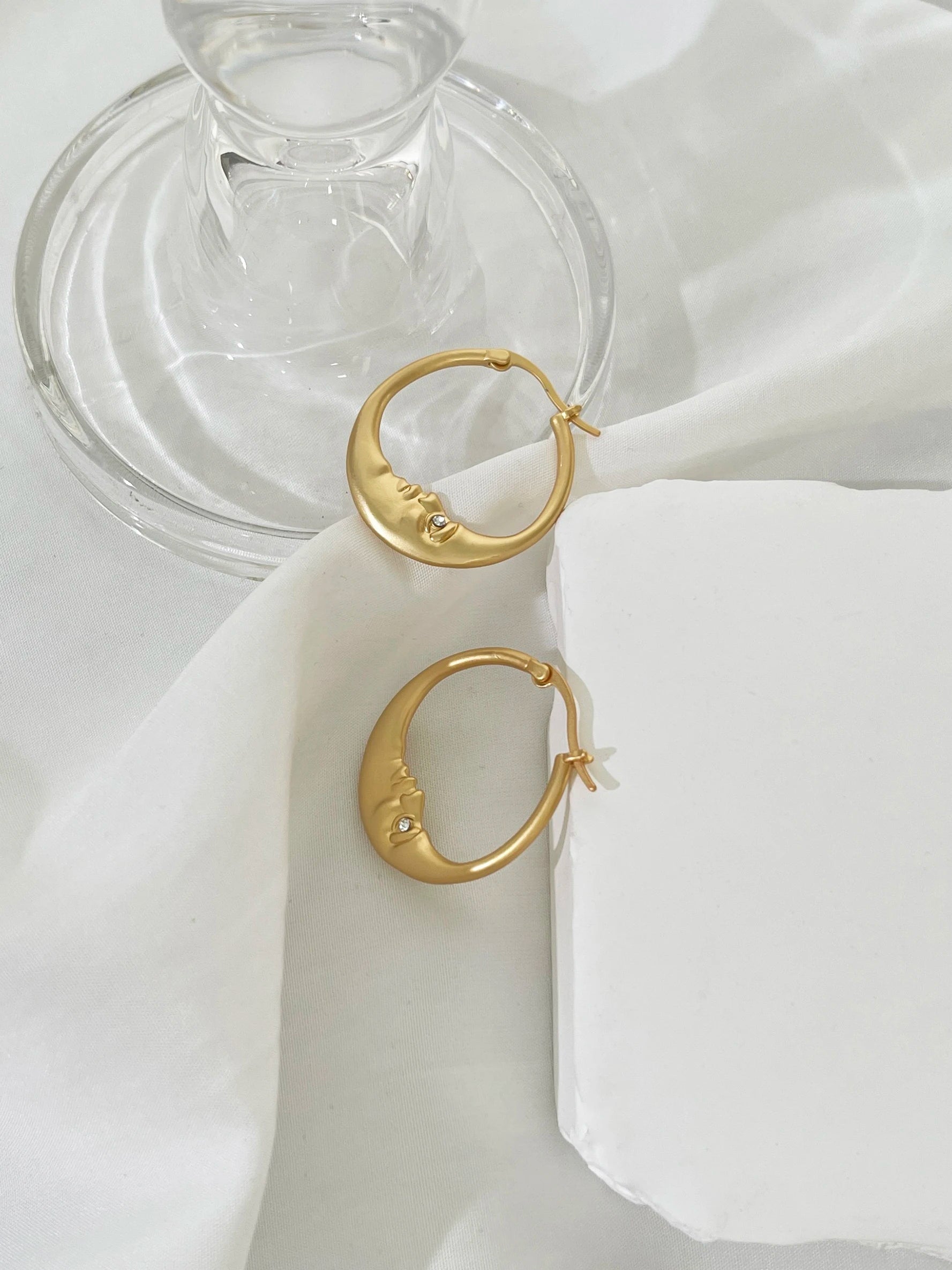 Gold hoop earrings on a white fabric with a glass dish in the background