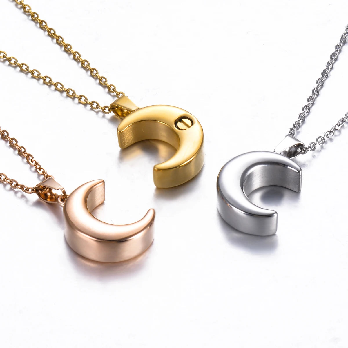Three crescent moon pendants on chains in gold, rose gold, and silver on a white background.