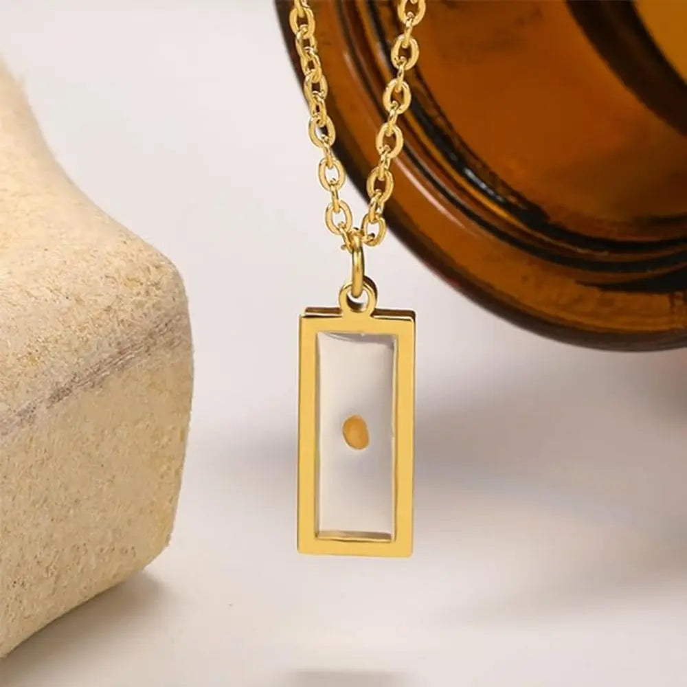 Gold necklace with a rectangular pendant featuring a small round object, set against a neutral background.