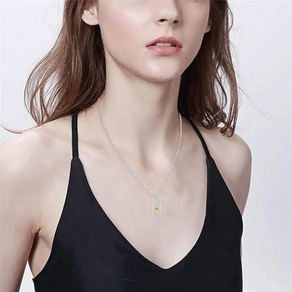 A close-up of a woman wearing a delicate silver chain necklace with a small rectangular pendant featuring a yellow flower detail, paired with a black camisole top against a light gray background.