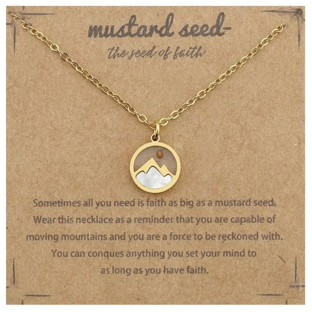 A gold-tone necklace with a circular pendant displayed on a brown kraft card. The pendant features a white inlay shaped like a mustard seed and small mountain-like shapes.