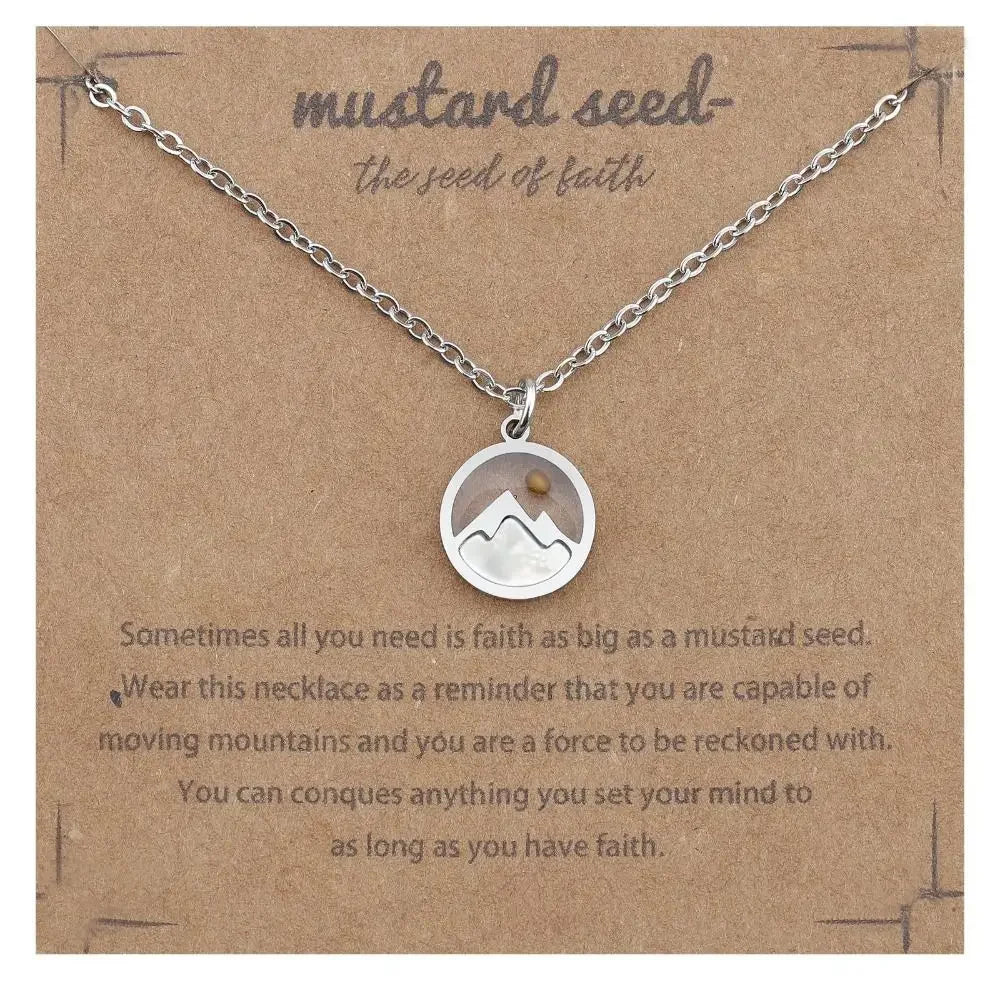 A silver necklace with a circular pendant featuring a mountain and sun design, displayed on a brown kraft card with the text 'mustard seed - the seed of faith' and a quote below.