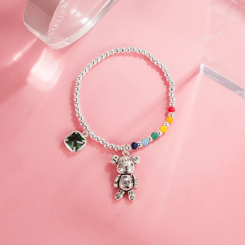 A silver beaded bracelet featuring a small teddy bear charm engraved with 'LOVE' and a green square gemstone charm, displayed on a pink background.
