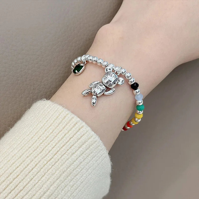 A close-up of a person's wrist wearing two beaded bracelets. One bracelet is silver with a small bear charm and a dark green oval stone.