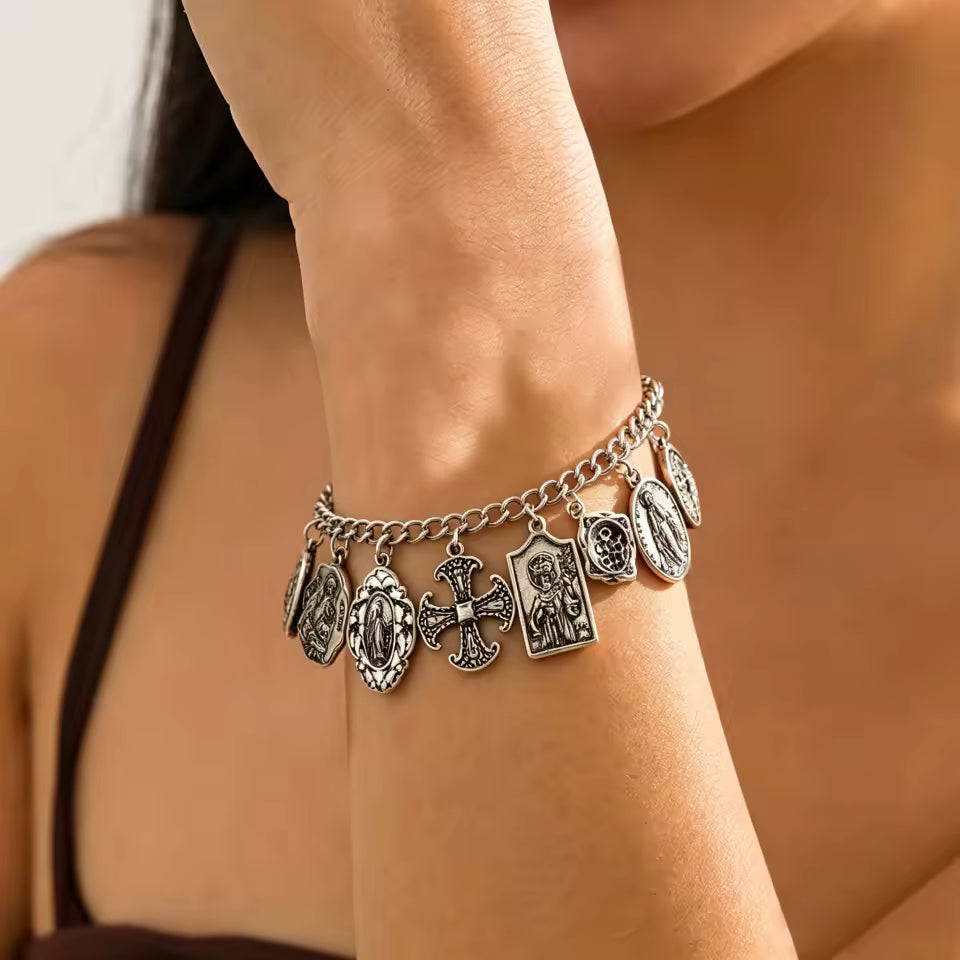 A close-up of a silver-toned charm bracelet worn on a woman's wrist. The bracelet features multiple religious saint and cross charms, including a central cross charm, shown against the model's skin.