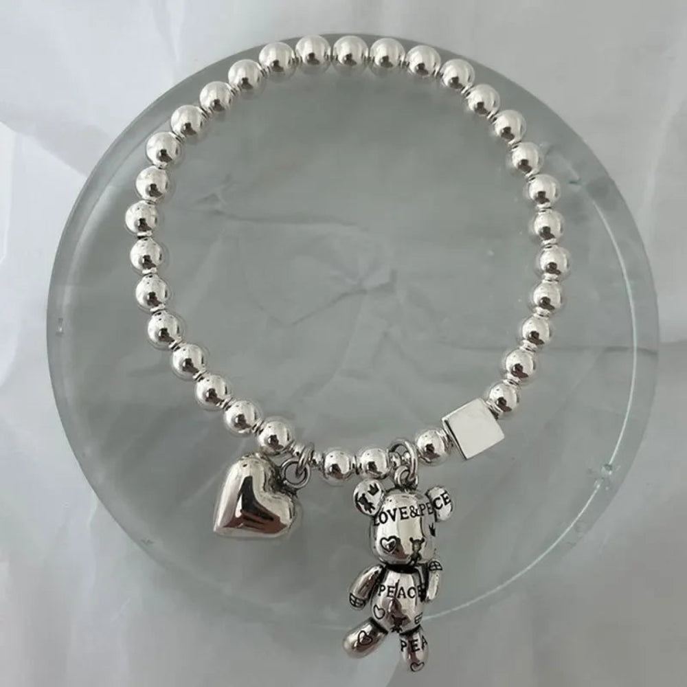 Silver beaded bracelet with teddy bear and heart charm on a clear stand.