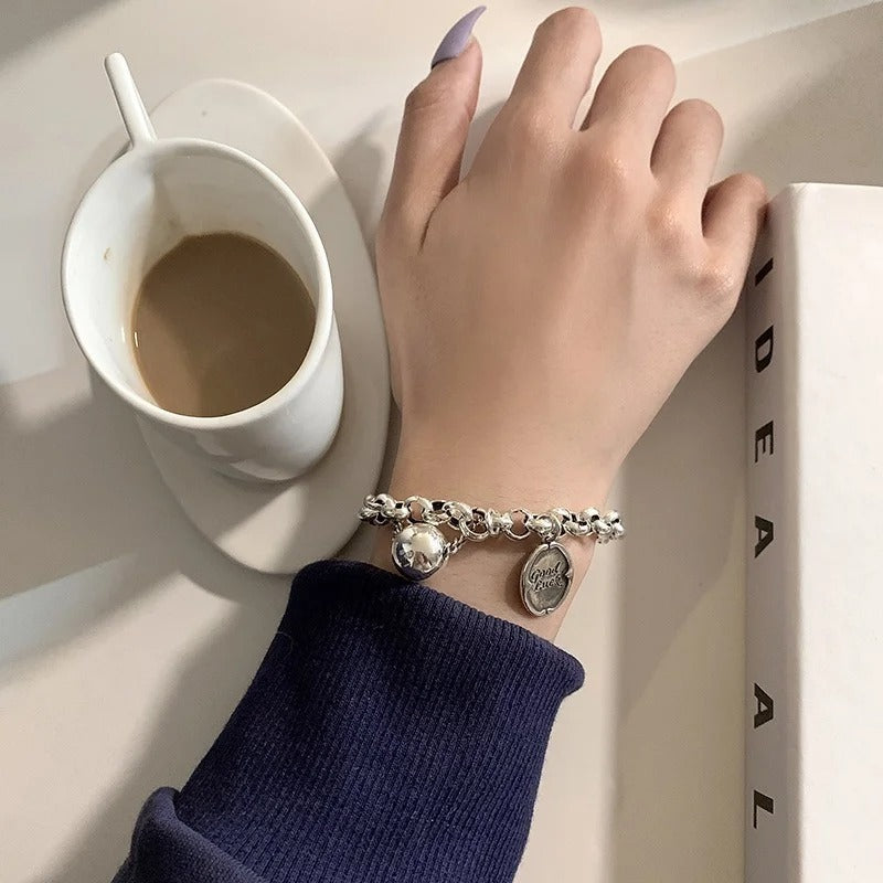 Hand wearing a silver bracelet with charms near a white cup of coffee on a light surface.