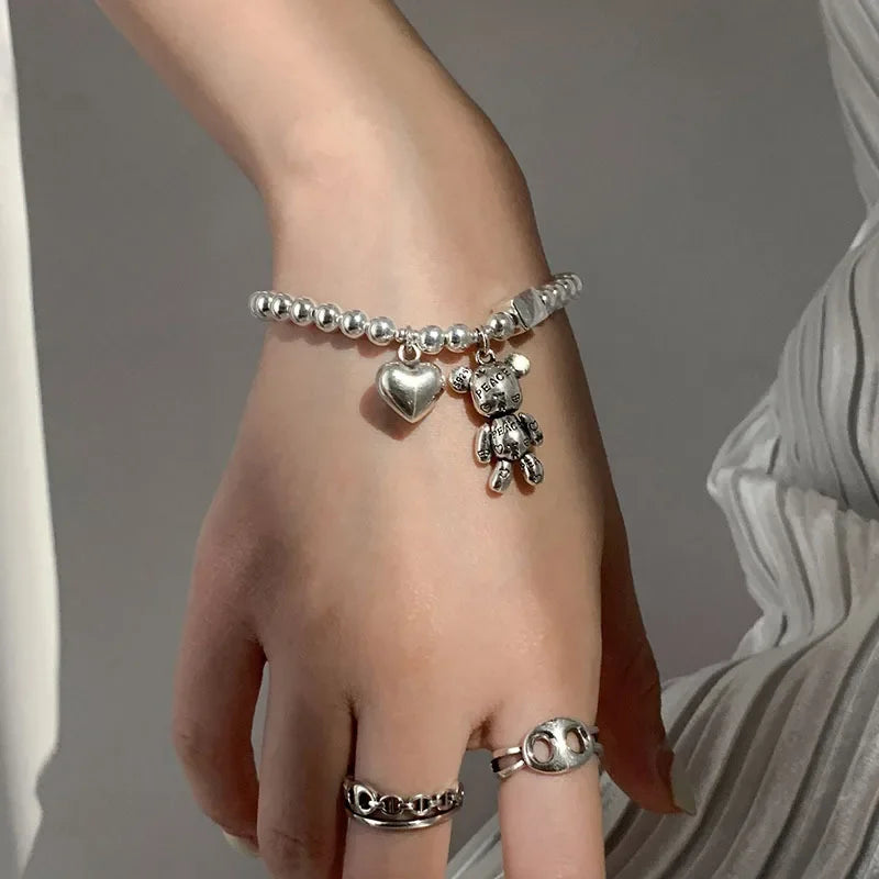 Hand wearing a silver bracelet with heart and teddy bear charms against a neutral background