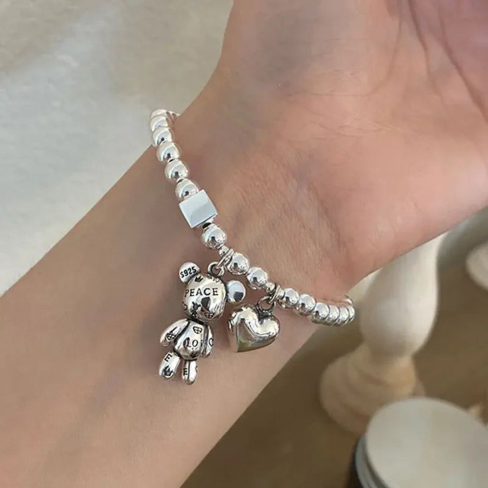 Silver bracelet with teddy bear and heart charms on a wrist against a neutral background