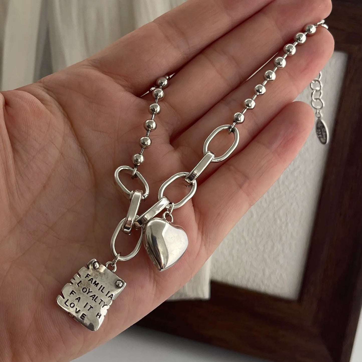 Silver necklace with heart and rectangular charm held in a hand against a neutral background